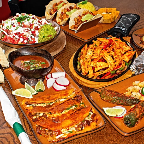 A table laden with various Mexican dishes, including nachos, tacos, fajitas, birria, and roasted peppers.